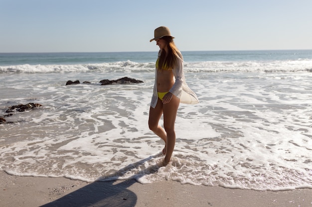 Bella donna in bikini e cappello che cammina sulla spiaggia al sole