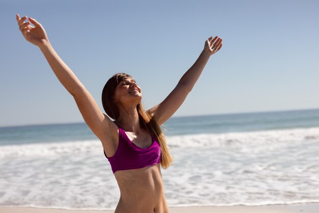 日差しの中でビーチに立って腕を持つビキニで美しい女性