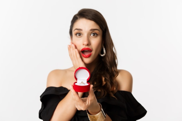 Beautiful woman in black dress making proposal, showing engagement ring and looking excited, standing over white background.