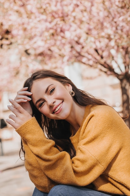 Beautiful woman in bright sweater posing on street against background of sakura. City portrait of attractive lady in yellow outfit smiling widely