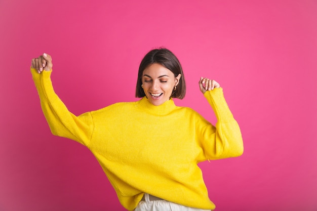 Красивая женщина в ярко-желтом свитере изолирована на розовой счастливой взволнованной танцующей подвижной улыбке
