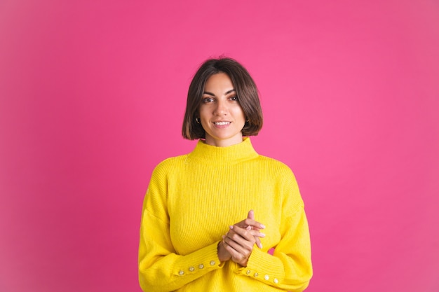 Красивая женщина в ярко-желтом свитере, изолированном на розовом, смотрит вперед с уверенной улыбкой