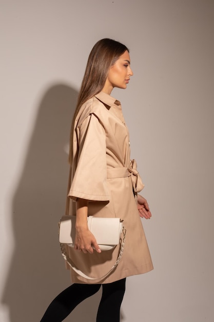 Красивая женщина в коричневом пальто, черных брюках, белых сапогах, держащая белую сумку, позирует перед камерой