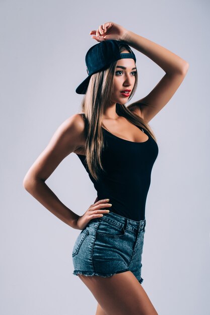 Beautiful woman in cap is having fun on a gray wall, smiling and looking to the side.