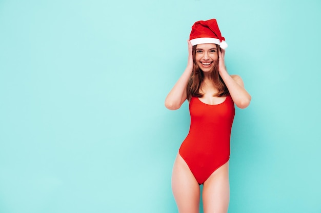 Beautiful woman celebrating New Year.Happy gorgeous female in red sexy body. She having fun at New Year's Eve party.Holiday celebration.Charming model in Santa Claus hat laughing near blue wall