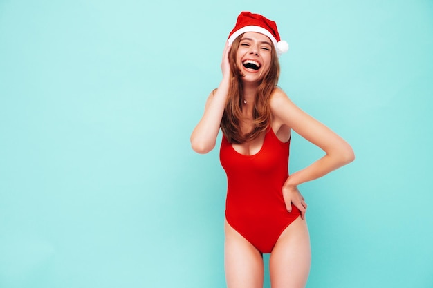 新年を祝う美しい女性。赤いセクシーな体で幸せなゴージャスな女性。彼女は大晦日のパーティーで楽しんでいます。休日のお祝い。青い壁の近くで笑っているサンタクロースの帽子の魅力的なモデル