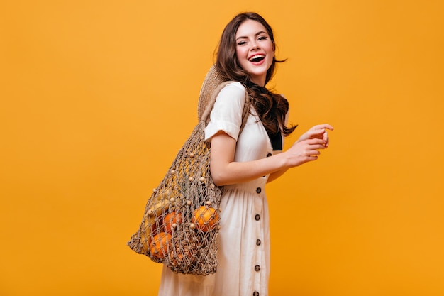 Beautiful woman in cotton dress laughs and holds string bag with fruits. Portrait of lady with wavy hair on orange background.
