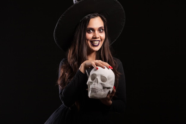 Beautiful woman dressed as witch for Halloween playing with a scary skull. Brunette witch doing magic with a skull.