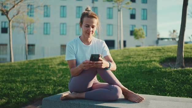 야외에서 스마트폰을 사용하여 소셜 미디어를 확인하는 운동복을 입은 아름다운 여성 도시 공원에서 쉬고 있는 젊은 요기 여성