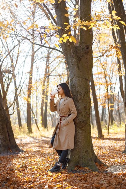 아름 다운 여자는 커피를 마시고 가을 공원에서 카메라를 위해 포즈를 취합니다. 커피와 함께 나무 근처에 서 있는 어린 소녀. 베이지색 코트를 입고 갈색 머리 여자입니다.