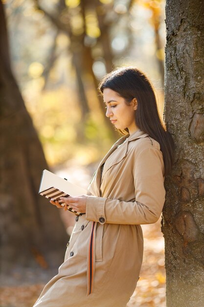 Beautiful woman drink coffee and posing for the camera in autumn park. Young girl standing near tree with a coffee. Brunette woman wearing beige coat.