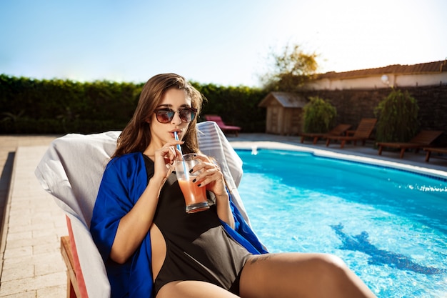 Beautiful woman drinking cocktail, lying on chaise near swimming pool