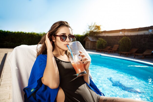 Beautiful woman drinking cocktail, lying on chaise near swimming pool