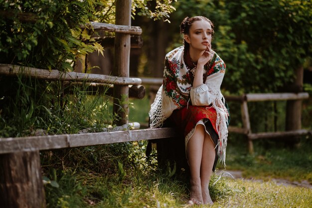 Beautiful woman in an embroidered traditional dress is sitting on the bench and looking into the distance