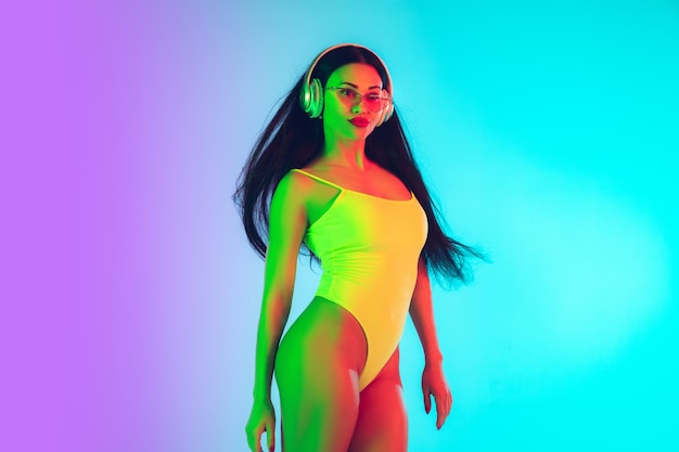 Beautiful woman in fashionable swimsuit isolated on gradient studio wall in neon light.