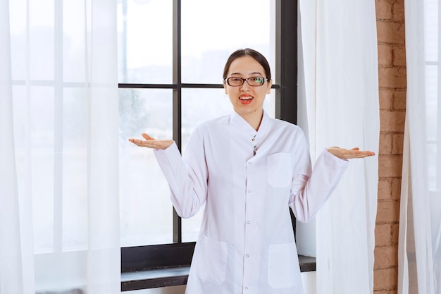 Beautiful woman in glasses and lab coat standing near window and shrugging shoulders .