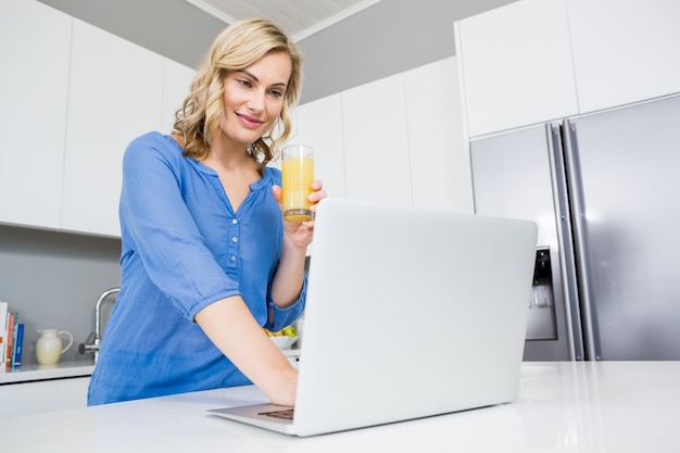 Beautiful woman holding a glass of juice using laptop in kitchen