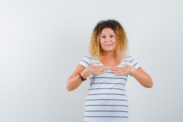 Beautiful woman holding hands on chest in t-shirt and looking surprised , front view.