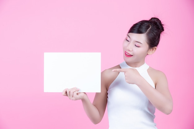 Beautiful woman holding a white board sheet on a pink background.