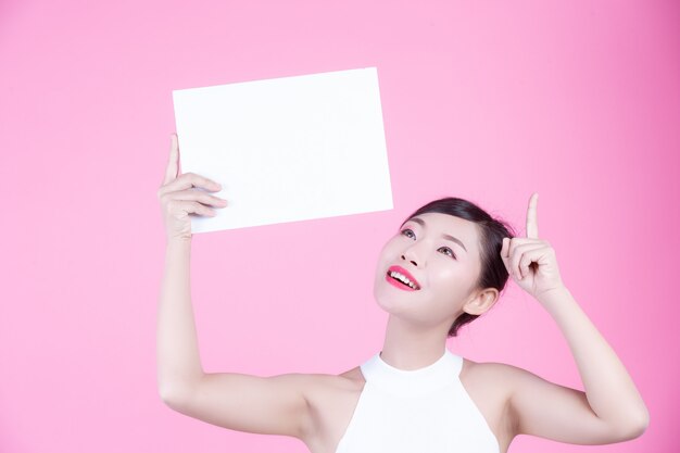 Beautiful woman holding a white board sheet on a pink background.