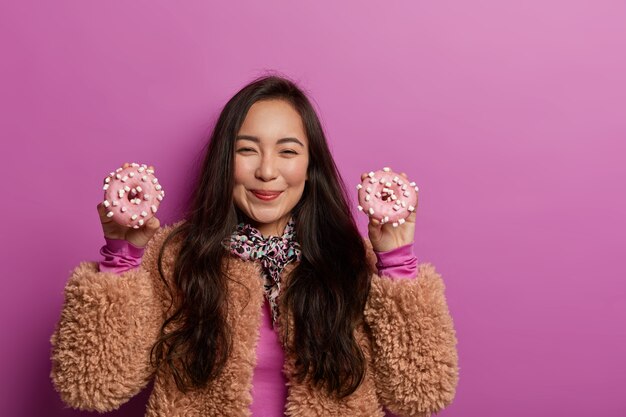 Beautiful woman holds two tasty donuts in both hands, has glad expression, feels temptation as keeps to diet, wears brown coat