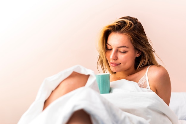 Beautiful woman laying in bed and looking down at a holding cup