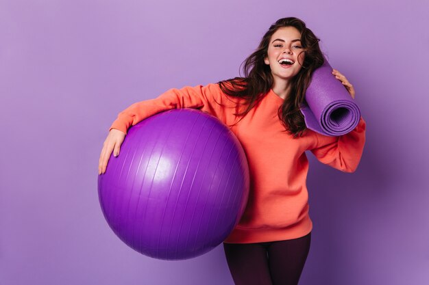 Beautiful woman in leggings and bright sweatshirt is smiling and posing with purple mat and fitball