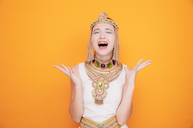 Beautiful woman like cleopatra in ancient egyptian costume angry and frustrated shouting and yelling raising arms with aggressive expression on orange
