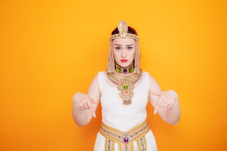 मुफ़्त फोटो beautiful woman like cleopatra in ancient egyptian costume confused pointing with index fingers down on orange