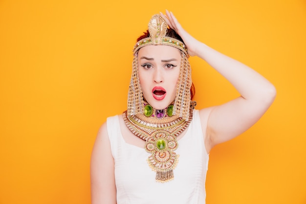 Beautiful woman like cleopatra in ancient egyptian costume confused and surprised holding hand on her head for mistake on orange