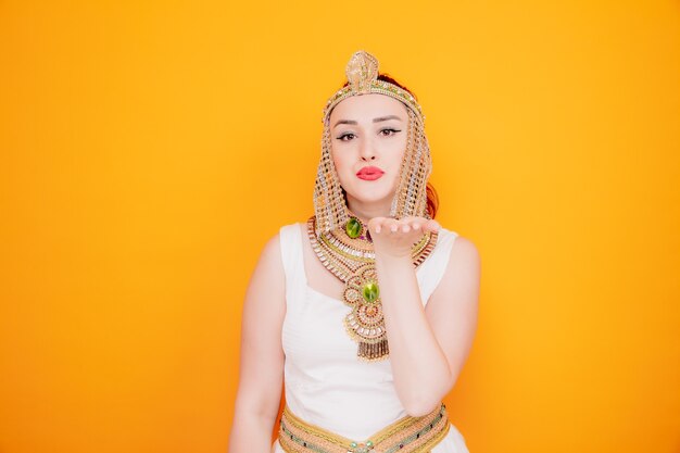 고대 이집트 의상을 입은 클레오파트라 같은 아름다운 여성이 오렌지색에 행복하고 긍정적인 공기 키스를 보낸다