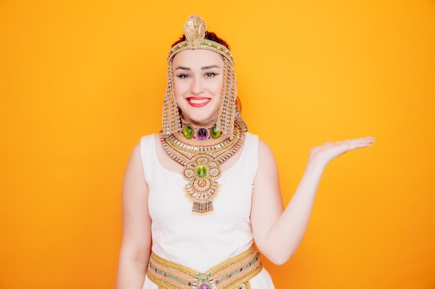 Bella donna come cleopatra in antico costume egiziano con un sorriso sul viso felice che presenta qualcosa con il braccio della mano sull'arancia