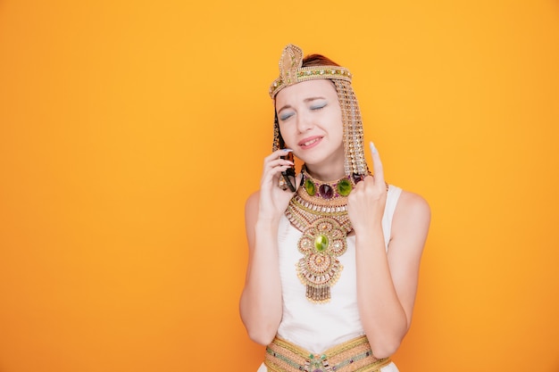 고대 이집트 의상을 입은 클레오파트라와 같은 아름다운 여성이 오렌지색에 실망한 표정으로 팔을 들어 올리는 휴대전화로 통화하는 동안 좌절한 표정을 하고 있다