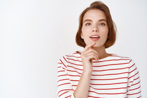 Beautiful woman looking thoughtful and sensual , touching lip with opened mouth, pondering something interesting, standing against white wall