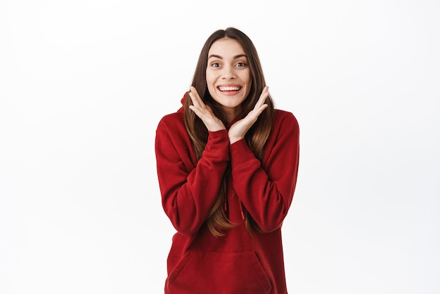 Beautiful woman looks surprised and happy hear amazing good news and rejoicing reaction of happiness to positive info standing in red hoodie against white background