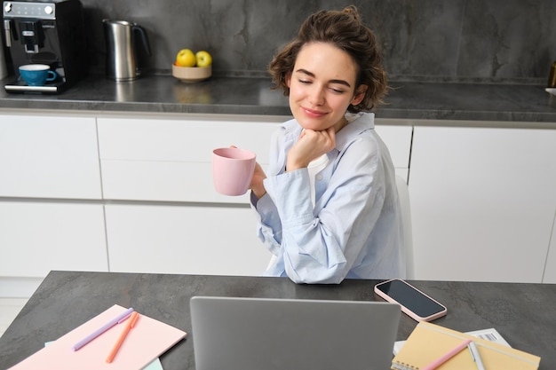 Beautiful woman managing her work from home drinking coffee and looking at laptop sitting in kitchen