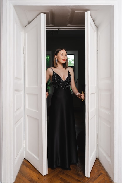 Free photo beautiful woman model dressed in an elegant long black dress in a fashion pose in a modern interior with white door on burgundy wall