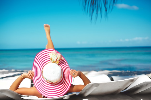 Beautiful woman model sunbathing on the beach chair in white bikini in colorful sunhat behind blue summer water ocean