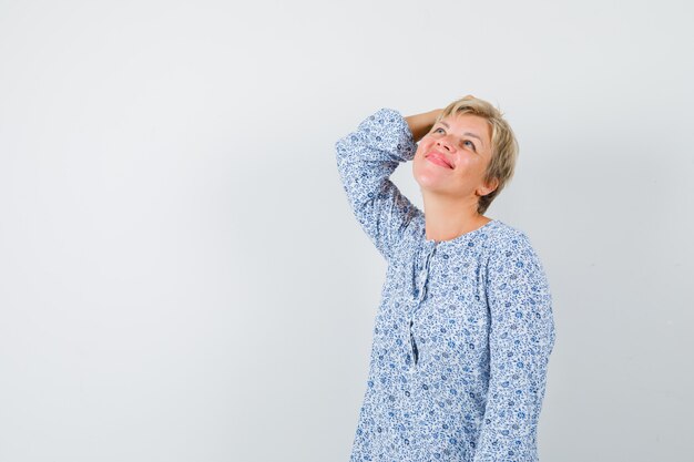 Beautiful woman in patterned blouse holding hand on head while looking up and looking happy , front view.