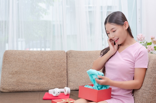 Beautiful woman in pink dress, sitting on the sofa, opening a gift box.