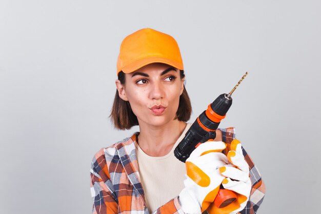 Beautiful woman in plaid shirt, hat and gloves, on gray wall holds drill to repair  home confident happy smile