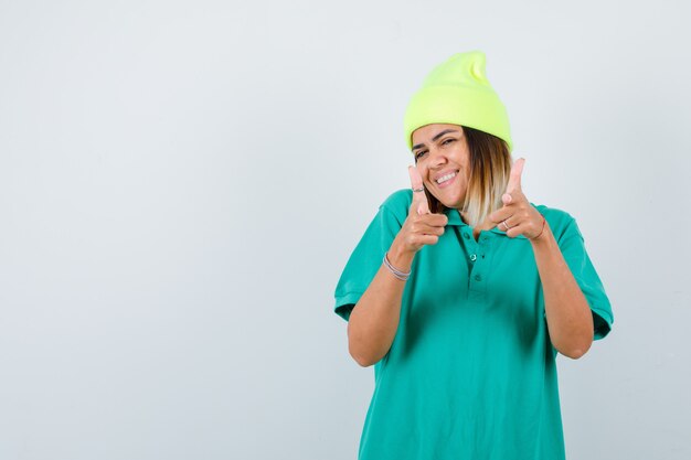 Beautiful woman pointing forward in polo t-shirt, beanie and looking happy. front view.