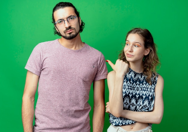 beautiful woman pointing at her displeased and frowning man , young couple man and woman over green wall