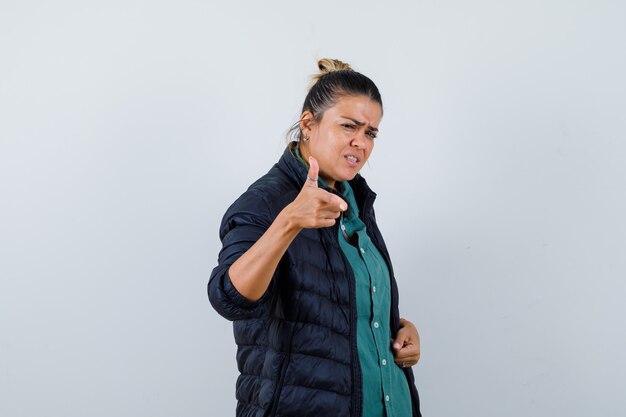 Beautiful woman pointing  with index finger in green shirt, black jacket and looking angry. front view.