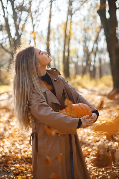 秋の公園で写真のポーズをとる美しい女性。カボチャを手に立って保持している若い女の子。ベージュのコートを着ているブロンドの女性。