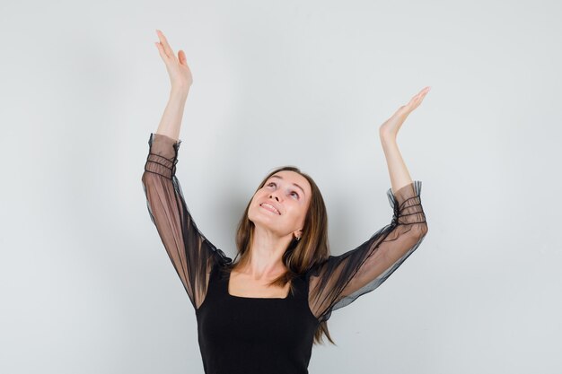 Beautiful woman raising arms with open palms while looking up in black blouse and looking merry 