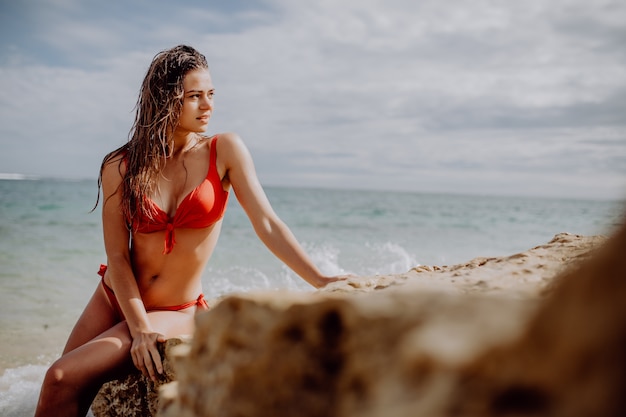 Beautiful woman in red bikini posing on beach sitting on rocks