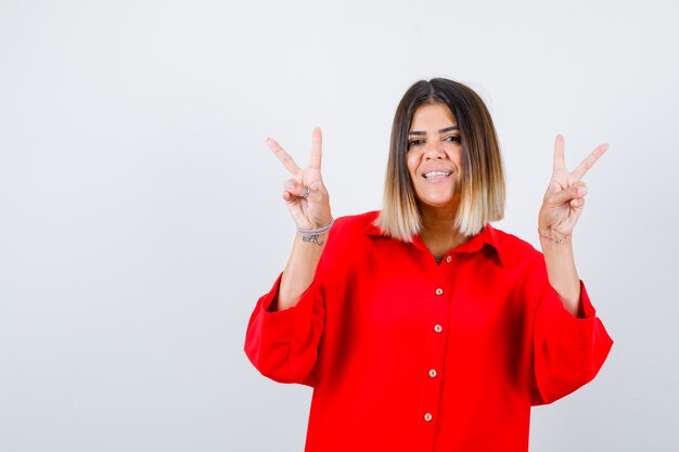 Beautiful woman in red blouse showing peace gesture and looking jolly , front view.