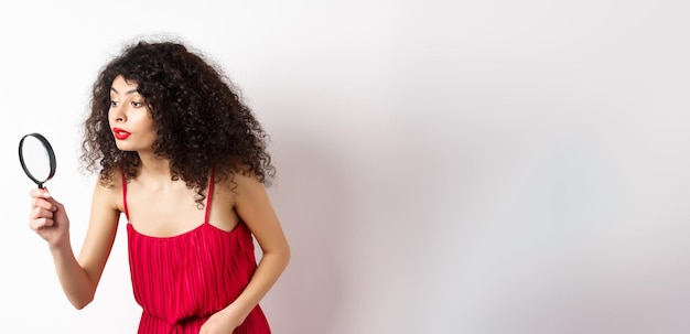 Beautiful woman in red dress searching for something looking aside through magnifying glass standing