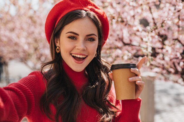 Beautiful woman in red outfit holds glass of tea and takes selfie on background of sakura. Portrait of brunette girl in hat smilimg and posing with coffee cup